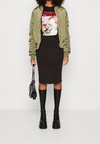 Olivově zelená bomber bunda, černé tričko s grafikou, přiléhavá černá midi sukně, černé kozačky na platformě po kolena a malá černá kabelka.