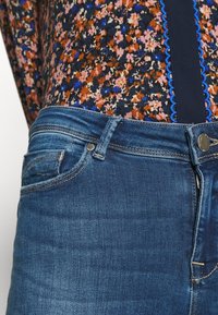 Jeans aus Denim in einem mittelblauen Waschung, mit klassischem Fünf-Taschen-Design, genähten Akzenten und einer glatten Textur. Darüber ein Hemd mit floralem Muster sichtbar.