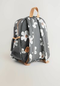 Grå ryggsäck med vitt blommönster och bruna detaljer. Har vadderade remmar, handtag på toppen och ett slätt, vattenavvisande material.