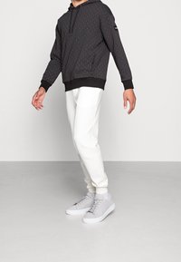 Sudadera negra con un patrón, con cordones, combinada con pantalones blancos ajustados y zapatillas grises. La sudadera cuenta con puños y dobladillo acanalados.