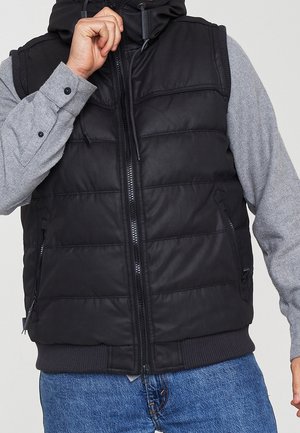 Schwarze gepolsterte Weste mit hohem Kragen, Reißverschlusstaschen und geripptem Saum; getragen über einem grauen Langarmshirt mit glatter Textur.