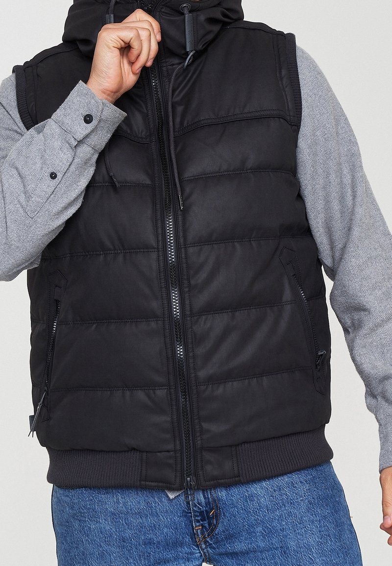 Gilet imbottito nero con colletto alto, tasche con zip e orlo a costine; indossato sopra una maglietta grigia a maniche lunghe, caratterizzata da una texture liscia.