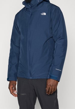 Man wearing a navy blue waterproof The North Face jacket with front zipper, adjustable cuffs, and zippered side pockets.