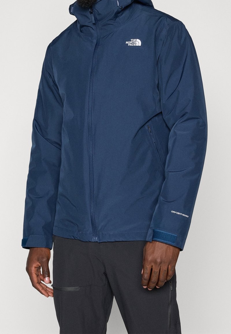 Homme portant une veste imperméable bleu marine The North Face avec fermeture éclair frontale, poignets réglables et poches latérales zippées.