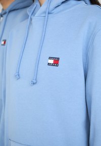 Hanorac bleu deschis cu șnur, având un mic logo rectangular în roșu, alb și bleu marin care spune "TOMMY JEANS" pe piept.