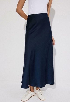 Žena v tmavomodrej dlhej sukni, bielom tielku bez rukávov a bielych šnurovacích teniskách stojí pred svetlým jednoliatym pozadím.