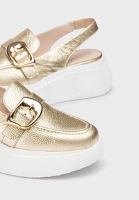 Chaussures à enfiler texturées en or avec une large sangle et une boucle dorée, dotées d'une semelle plate blanche. L'arrière présente un design à talon ouvert.