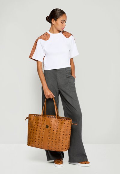 Woman wearing white cropped top with brown accents, gray wide-leg trousers, holding large brown tote bag with monogram pattern.