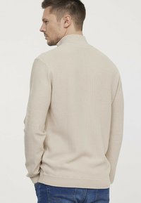 Pull en tricot beige avec un motif texturé, col haut et poignets et ourlet côtelés. Porté avec un jean bleu, vu de dos.