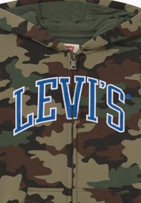Camouflage lynlås-hættetrøje i brun, grøn og sort med en blå broderet "LEVI'S"-logo på brystet og en frontlomme. Blødt stof.