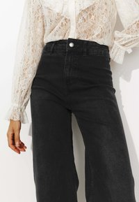 Jean noir taille haute avec une coupe droite, dotés d'une fermeture par bouton sur le devant et d'une texture subtile, associés à un blouse blanche en dentelle.