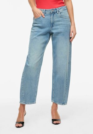 Femme portant un jean bleu clair à jambes droites avec cinq poches, associé à des talons ouverts noirs et un haut rouge sans manches, debout sur fond blanc.