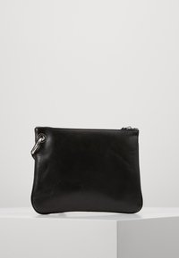 Pochette en cuir noir avec fermeture éclair argentée et détail en métal, présentée sur un cube blanc sur fond blanc.