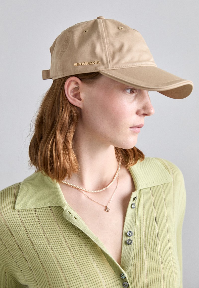 Beige baseball cap with a curved brim, made of soft fabric, featuring a gold logo on the side. Worn with a green, ribbed collared top.