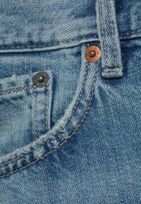 Gros plan sur une poche de jean bleu en denim avec rivets, passant de ceinture et détails visibles de couture.