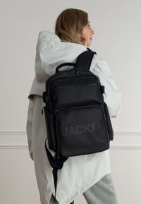 Zaino in nylon nero con più scomparti, chiusure a zip e un logo prominente. Il design include spallacci imbottiti per il comfort.
