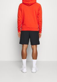 Felpa rossa con cappuccio, dotata di tasca a marsupio e orlo a coste, abbinata a pantaloni corti neri. Calzini e scarpe bianchi completano l'outfit.