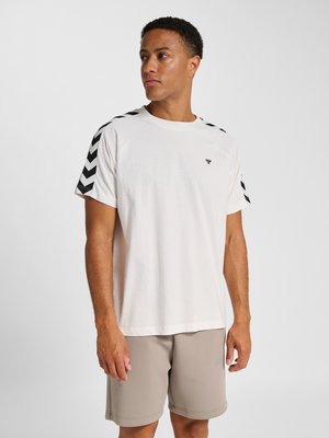 Mand iført hvid T-shirt med korte ærmer med sorte chevronstriber på skuldrene og beige shorts, der kigger til venstre.