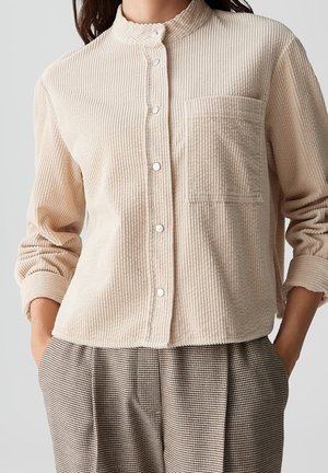 Chemise en velours côtelé beige avec un col mao, des boutons sur le devant et une poche poitrine, associée à un pantalon à carreaux aux tons terreux.