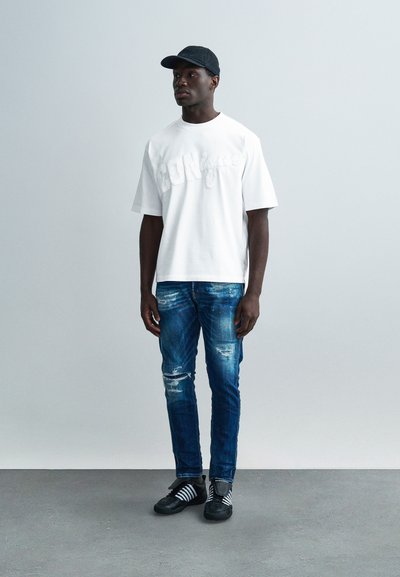 Λευκό T-shirt με κοντά μανίκια και ανάγλυφο κείμενο, τζιν παντελόνι σε φθαρμένο μπλε χρώμα και μαύρα παπούτσια με λευκές ρίγες. Φοράει μαύρο καπέλο.