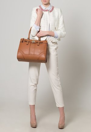 Femme vêtue d'un pantalon blanc sur mesure, d'une veste blanche texturée avec un col coloré, tenant un grand sac à main en cuir marron.