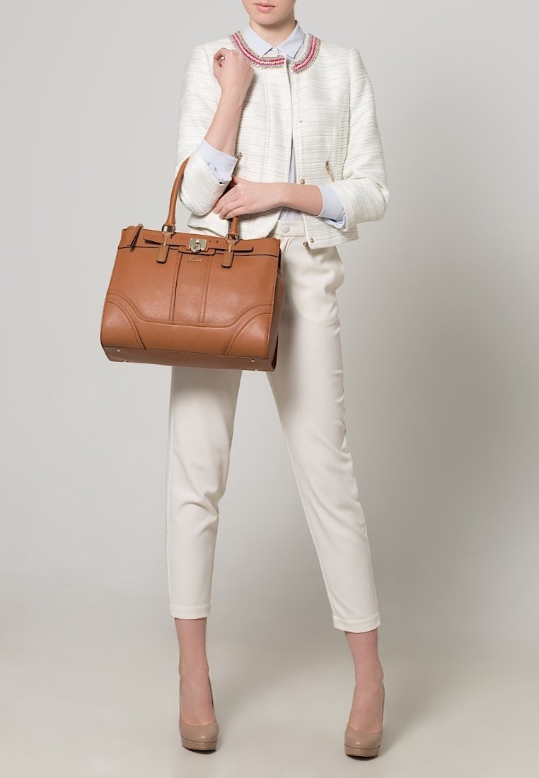Femme vêtue d'un pantalon blanc sur mesure, d'une veste blanche texturée avec un col coloré, tenant un grand sac à main en cuir marron.