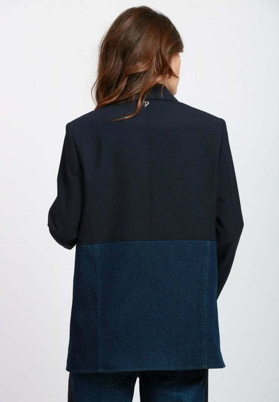 Blazer scuro dal design strutturato, con parte superiore nera uniforme e sezione inferiore in denim scuro e testurizzato. Sul retro è presente un dettaglio logo discreto.