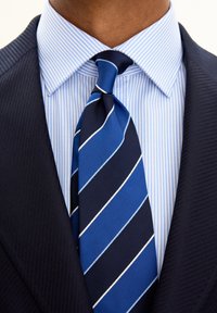 Giacca da abito blu navy con tessuto strutturato, camicia a righe azzurra chiara e cravatta blu spessa con strette righe bianche. Vista ravvicinata.