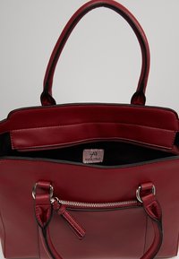 Czerwona torba typu tote z ekoskóry z dwoma uchwytami, kieszenią na zamek, gładką fakturą i czarnym wnętrzem. Widoczna metka z logo wewnątrz.