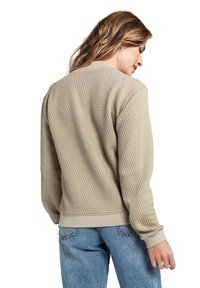 Beige strukturierter Pullover mit einem Waffelstrickdesign, gerippten Bündchen und Saum. Das Modell trägt ihn mit hellblauen Jeans.