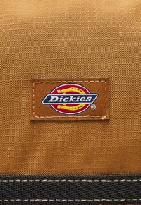 Étiquette en tissu marron affichant le logo "Dickies" en bleu et jaune, brodée sur un fond texturé orange avec des accents noirs.