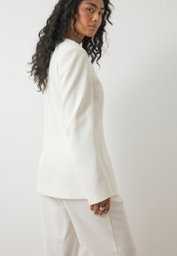 Valkoinen bleiseri, jossa on strukturoitu muotoilu ja pitkät hihat, paritettu yhteensopivien housujen kanssa. Sileä pinta ja kevyt kiilto. Tyylikäs siluetti.