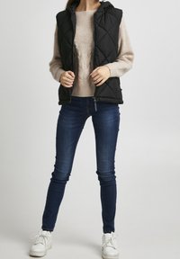 Czarna pikowana kamizelka z kapturem, zestawiona z beżowym swetrem z kablem i niebieskimi obcisłymi dżinsami. Model nosi białe sneakersy.