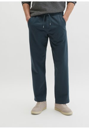 Mann trägt dunkelblaue Hose mit Kordelzug, Hände in den Taschen, beigefarbene Sneaker und ein schwarz-weiß gestreiftes Langarmshirt.