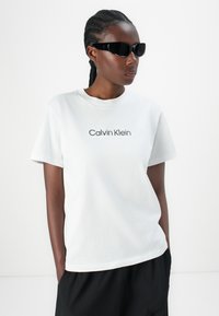 T-shirt blanca de algodón de Calvin Klein con mangas cortas y escote redondo; presenta texto del logo en negro. Combinado con gafas de sol negras.