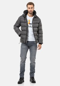 Bauchtasche aus mattem Dunkelgrau, mit einem Reißverschluss und einer verstellbaren Kapuze. Trägt ein weißes T-Shirt mit einem bunten Grafikdesign, kombiniert mit grauen Jeans und schwarzen Stiefeln.