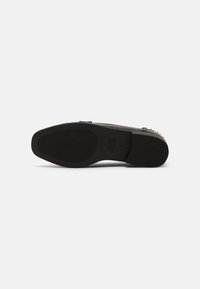 Semelle d'une chaussure plate noire à enfiler, avec des motifs de grip texturés et des détails en clous dorés sur le bord supérieur, présentée sur fond blanc.