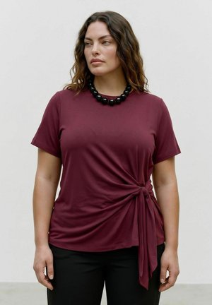 Femme portant un haut bordeaux à manches courtes avec un nœud sur le côté et un collier de perles noires, debout devant un fond clair uni.