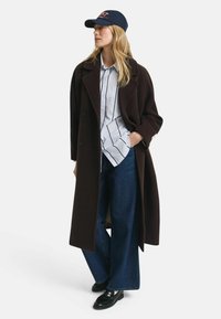 Long brown overcoat, striped button-up shirt, wide blue jeans, and black loafers. Finished with a navy cap featuring red text.