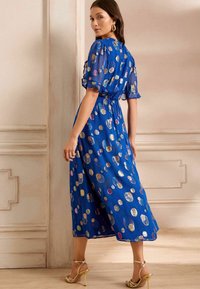 Modra midi obleka s naborki, ki ima barvite krožne vzorce. Prosojna tkanina s poudarjeno pasom in tekočo krilo. Zlate sandale.