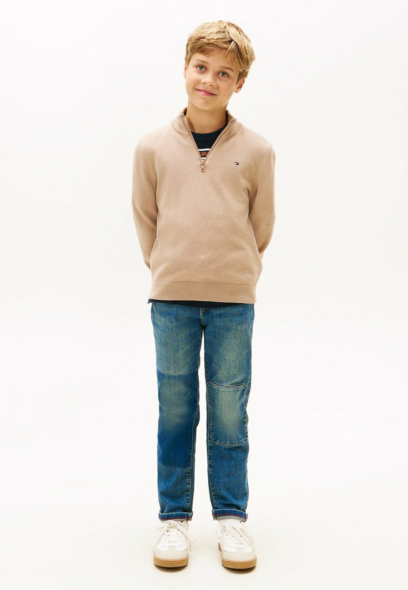 Blonde jongen staat met zijn handen achter zijn rug, draagt een beige ziptrui, blauwe jeans met patches en witte sneakers op een witte achtergrond.
