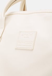 Sac de couleur crème avec une étiquette carrée cousue portant l'inscription "anello", avec un texte en relief sur le design et l'utilisation toute saison.