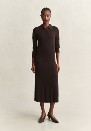 Femme debout portant une robe en maille côtelée marron foncé à manches longues avec un col boutonné et des chaussures à talons bas assorties.