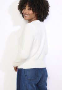 Bílý nadýchaný svetr se zaobleným spodním lemem, zdobený jemnými detaily. Kombinován s modrými denimovými džíny.