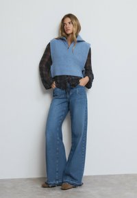 Chaleco de punto azul claro con un cuello alto sobre una camisa de cuadros oscuros de manga larga, combinado con unos jeans de mezclilla azul de pierna ancha y zapatos marrones de punta.