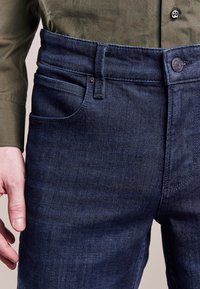 Man som bär mörkblå jeans och en olivgrön långärmad skjorta, med handen avslappnad vid sin sida.