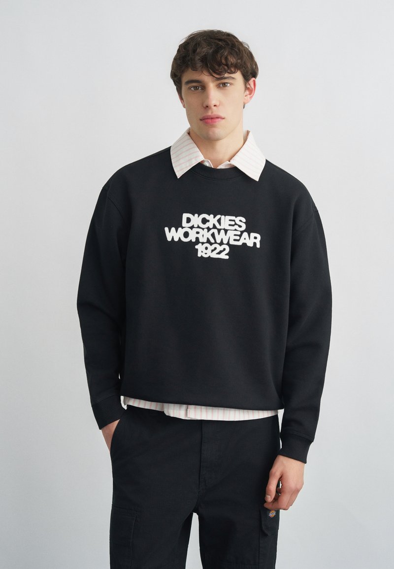 Giovane uomo con capelli corti e ricci che indossa una felpa nera Dickies Workwear 1922 sopra una camicia bianca con colletto, in piedi con una mano nella tasca.