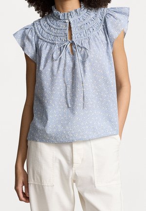 Mujer con blusa sin mangas azul claro con estampado floral, cuello con volantes y detalle de lazo, combinada con pantalones blancos de cintura alta.