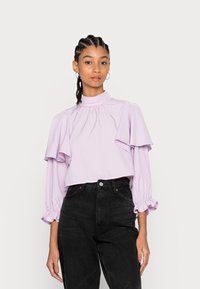 Blusa cropped em lavanda com mangas flare e detalhes na gola franzida. Combinada com jeans pretos de cintura alta. Textura suave, design casual.