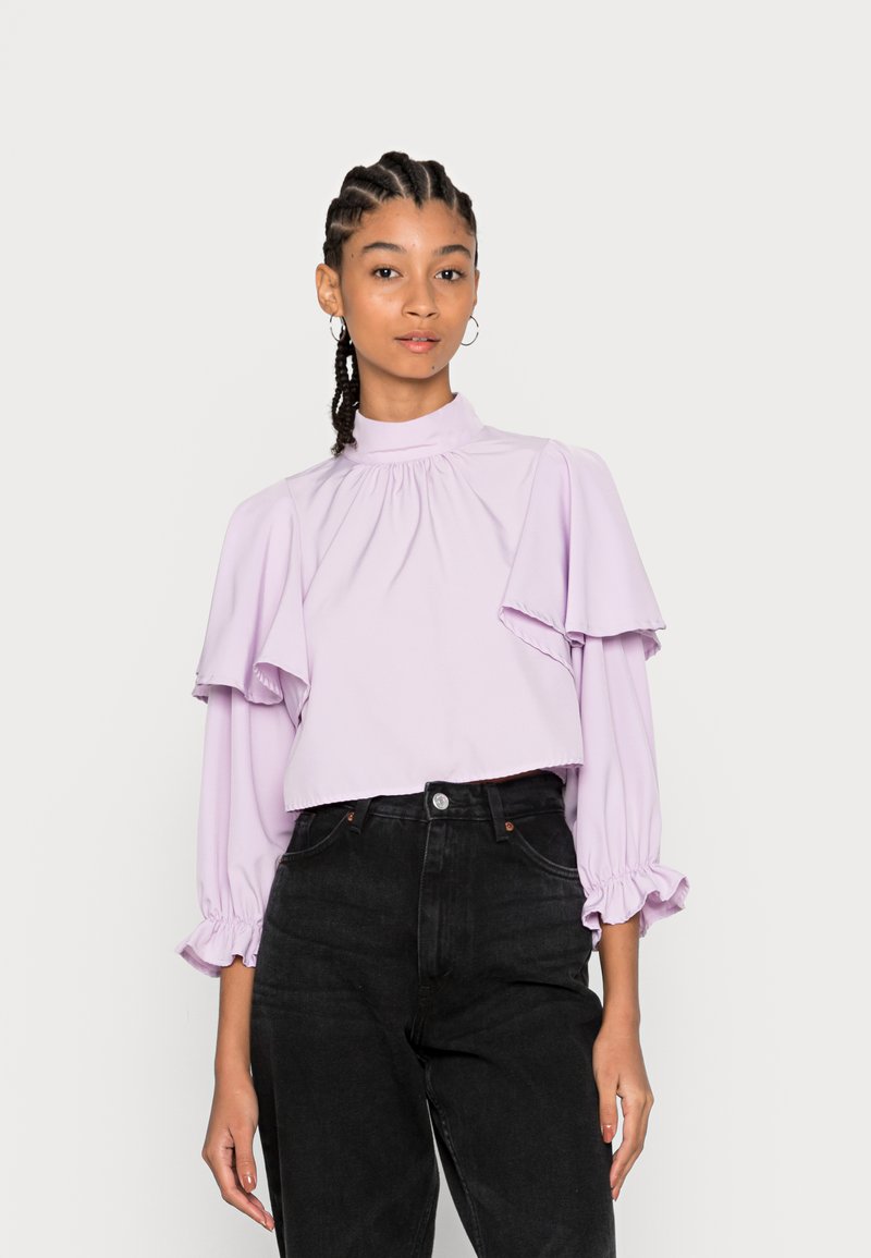 Blusa cropped em lavanda com mangas flare e detalhes na gola franzida. Combinada com jeans pretos de cintura alta. Textura suave, design casual.